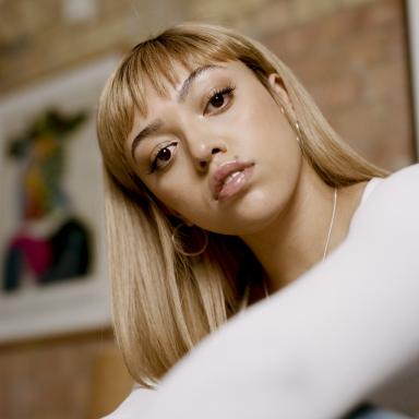 Woman with long blonde hair and fringe wearing white top and jeans looks at camera